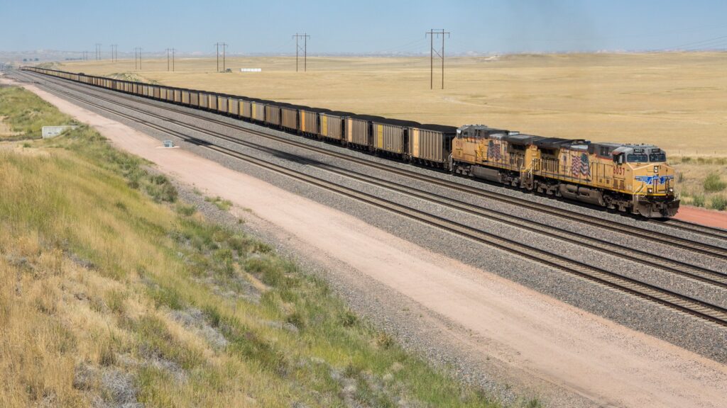 A freight train with multiple cargo cars travels along tracks in a rural landscape, with open fields and power lines in the background.