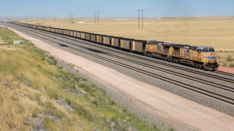A freight train with multiple cargo cars travels along tracks in a rural landscape, with open fields and power lines in the background.