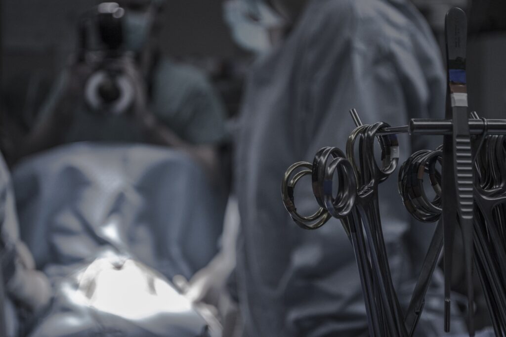 A surgical scene showing medical instruments on a tray, with a medical professional in surgical attire and another person in the background holding a camera.