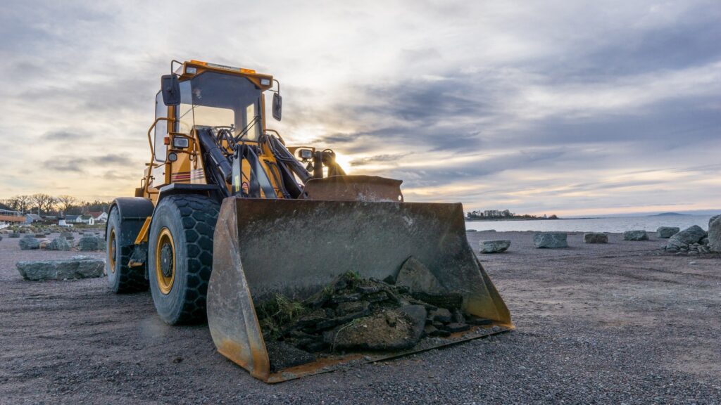 A front-end loader parked on a gravel area, with rocks and a scenic lakeside view in the background under a cloudy sky.