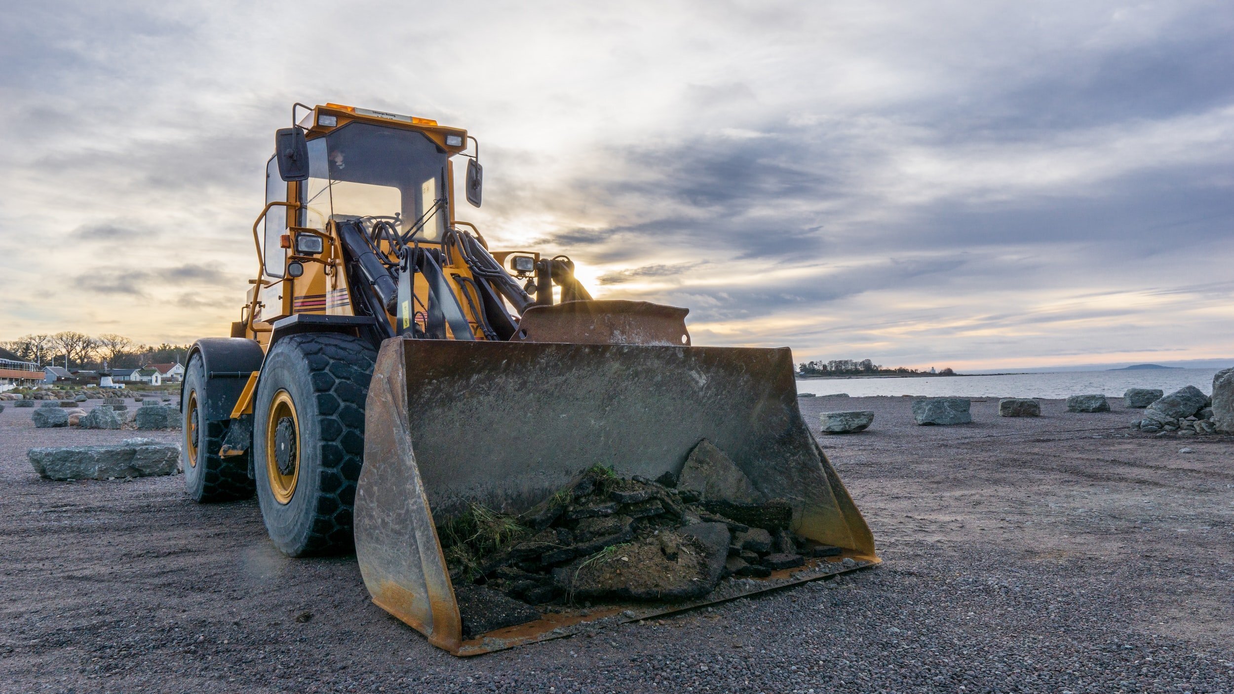 A front-end loader parked on a gravel area, with rocks and a scenic lakeside view in the background under a cloudy sky.