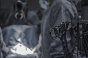 A surgical scene showing medical instruments on a tray, with a medical professional in surgical attire and another person in the background holding a camera.