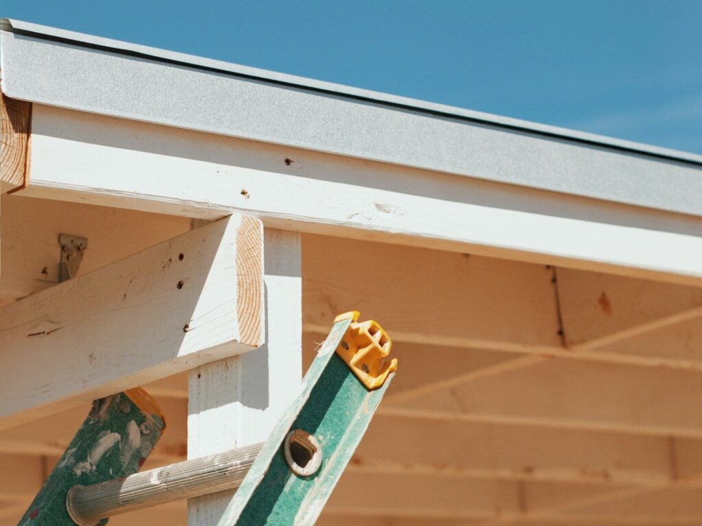 A green ladder leaning against a wooden structure with a partially visible roof under a clear blue sky.
