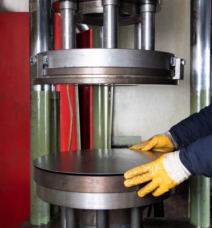 A person wearing yellow gloves operates a hydraulic press with a circular metal plate beneath it in a workshop setting.