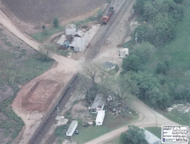 An aerial view of a rural area featuring railroad tracks, silos, various buildings, and scattered debris.